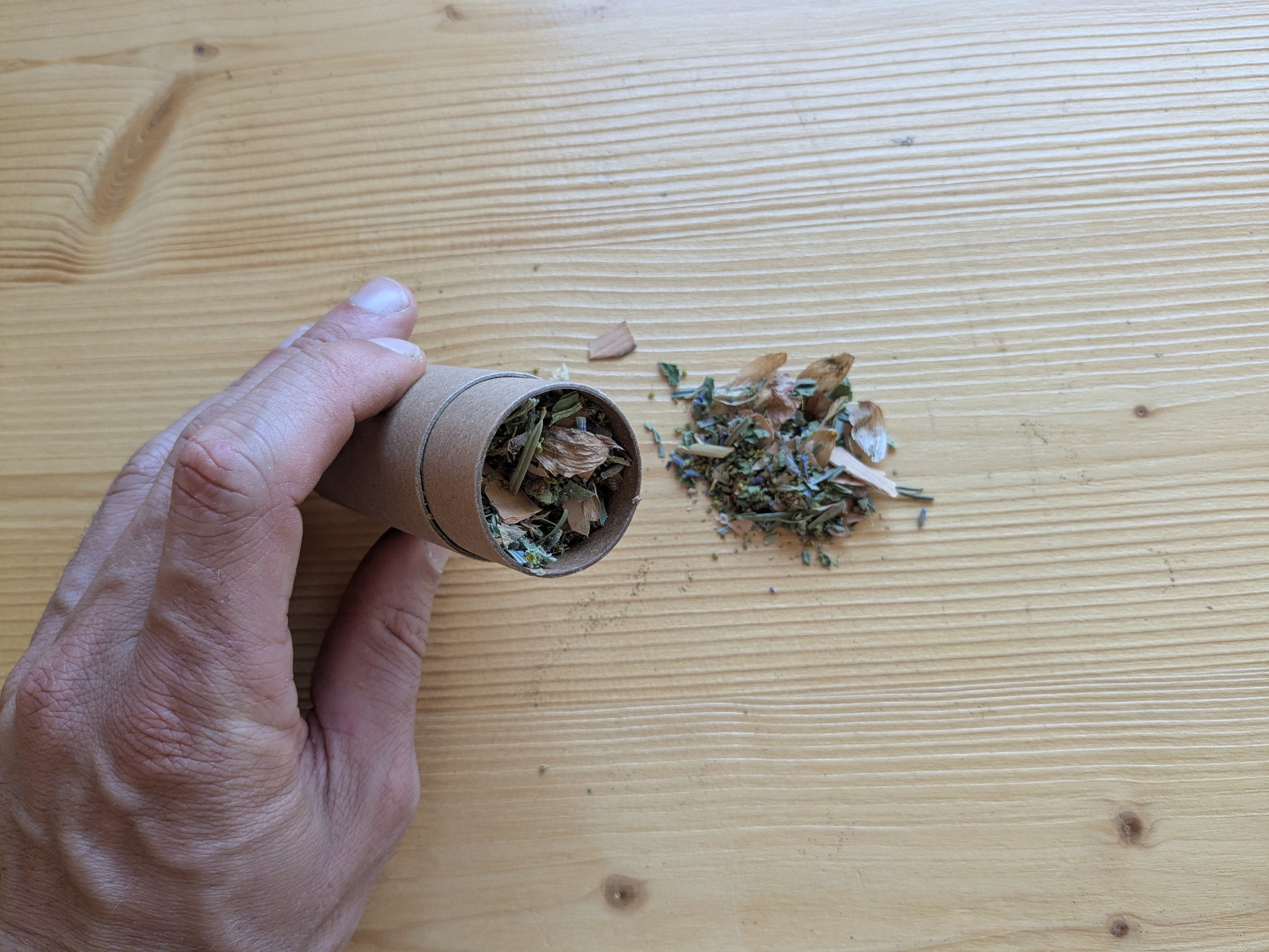 Hand hält Kartondose und schüttet Räuchermischung aus Kräutern auf hellen Holztisch