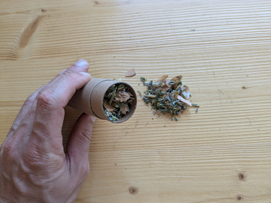 Hand hält Kartondose und schüttet Räuchermischung aus Kräutern auf hellen Holztisch