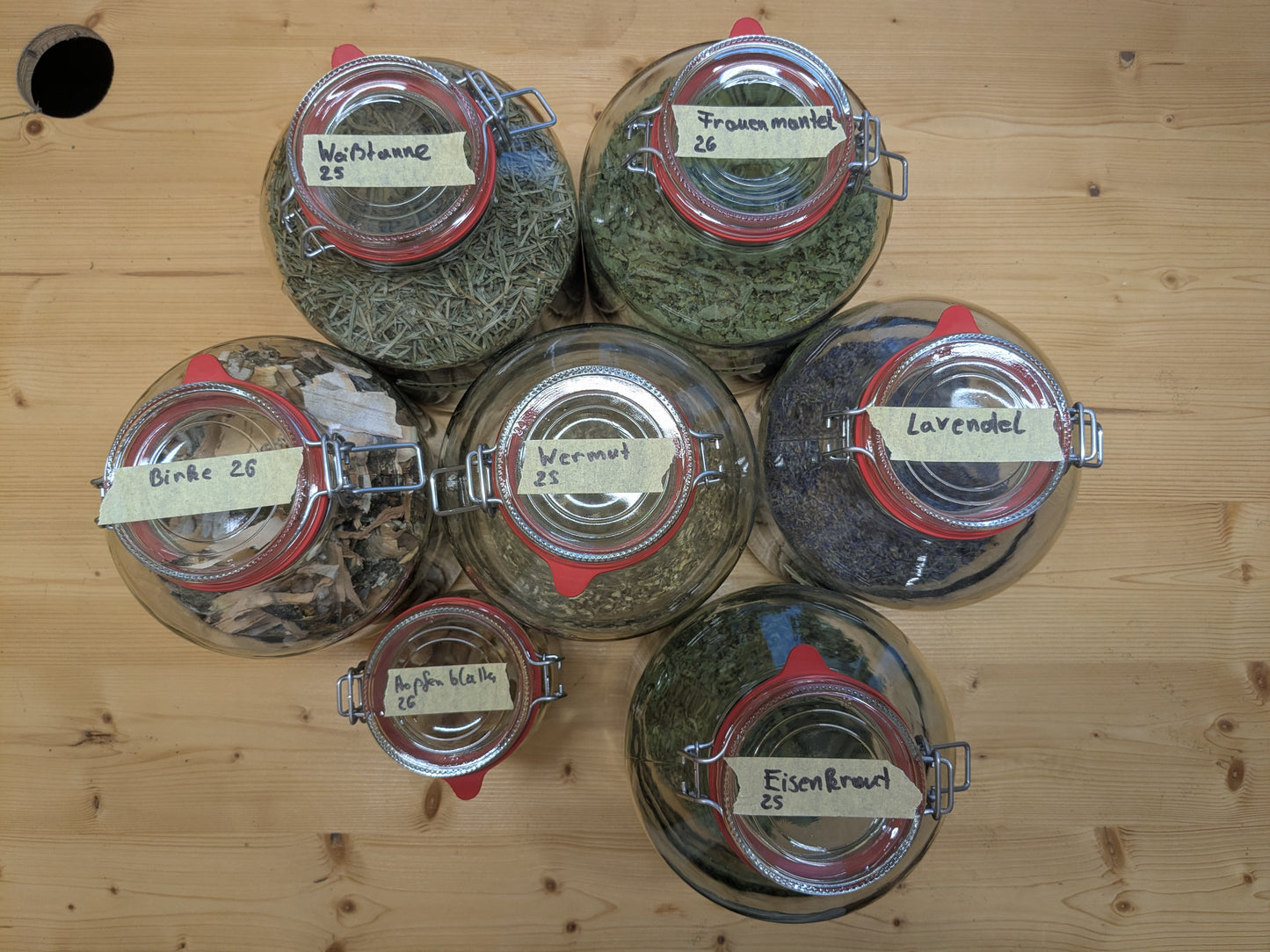 Draufsicht auf mehrere Bügelgläser mit beschrifteten Kräutern wie Lavendel, Wermut und Hopfenblüten auf Holztisch