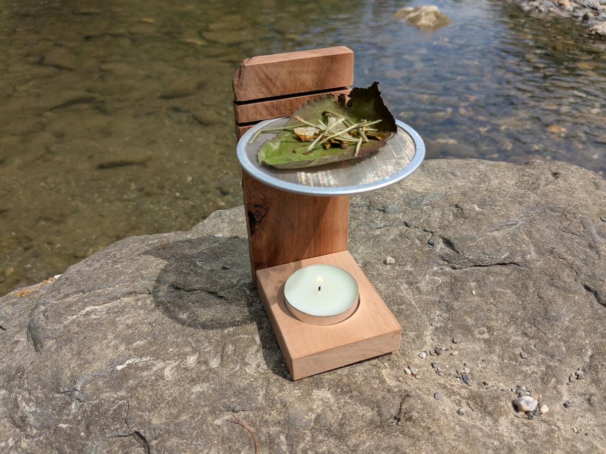 Räucherstövchen mit Kräutern auf Sieb und Teelicht in natürlicher Umgebung am Fluss.